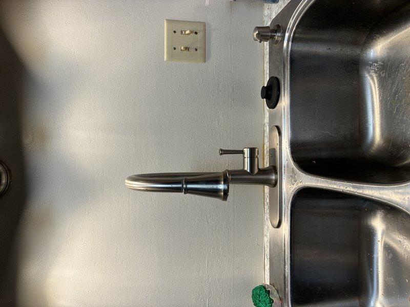 Installed customer-supplied single-handle kitchen faucet on double-basin stainless steel sink. Completed mounting and connection of the modern tall-spout faucet with black accent handle, ensuring proper positioning between basins and verifying all supply line connections for leak-free operation.