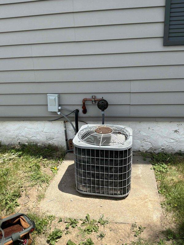 Provided estimate for temporary disconnect and reconnect of outdoor air conditioning condensing unit to accommodate landscaping work being performed at the property. The existing AC system features an older model condenser with copper refrigerant lines, wall-mounted shut-off valves, and electrical disconnect box mounted on the building foundation. Unit is positioned on a concrete pad adjacent to white vinyl siding with standard residential HVAC configuration showing typical outdoor weathering and patina from exposure to elements.