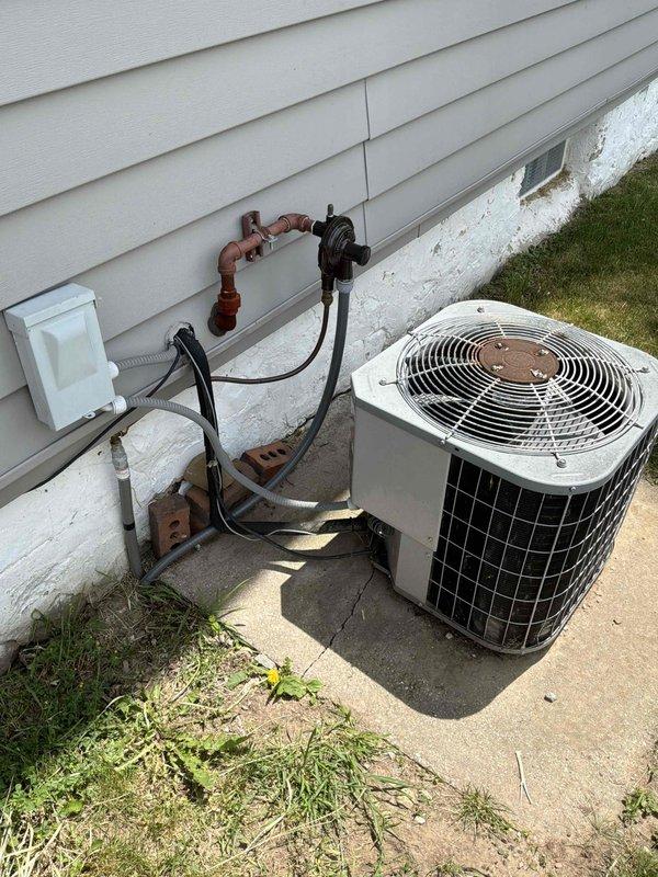 Provided estimate for temporary disconnect and reconnect of outdoor air conditioning condensing unit to accommodate landscaping work being performed at the property. The existing AC system features an older model condenser with copper refrigerant lines, wall-mounted shut-off valves, and electrical disconnect box mounted on the building foundation. Unit is positioned on a concrete pad adjacent to white vinyl siding with standard residential HVAC configuration showing typical outdoor weathering and patina from exposure to elements.