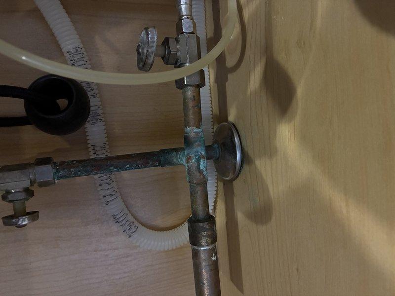 Responded to service call for diagnosis of slow leak under kitchen sink. Upon arrival, conducted thorough inspection of under-sink plumbing assembly. Identified significant corrosion and verdigris buildup on copper/brass pipe fittings at T-junction connection, indicating prolonged moisture exposure as source of leak. Corroded fittings had compromised seal integrity, allowing water to seep through connections. Documented existing double-basin stainless steel sink installation with single-handle chrome faucet for service records. Assessed condition of flexible supply lines and drain assembly. Provided customer with diagnostic findings and recommendations for repair of corroded fittings to resolve active leak issue. Customer also expressed interest in future installation of additional sink fixture, which was noted for follow-up consultation.