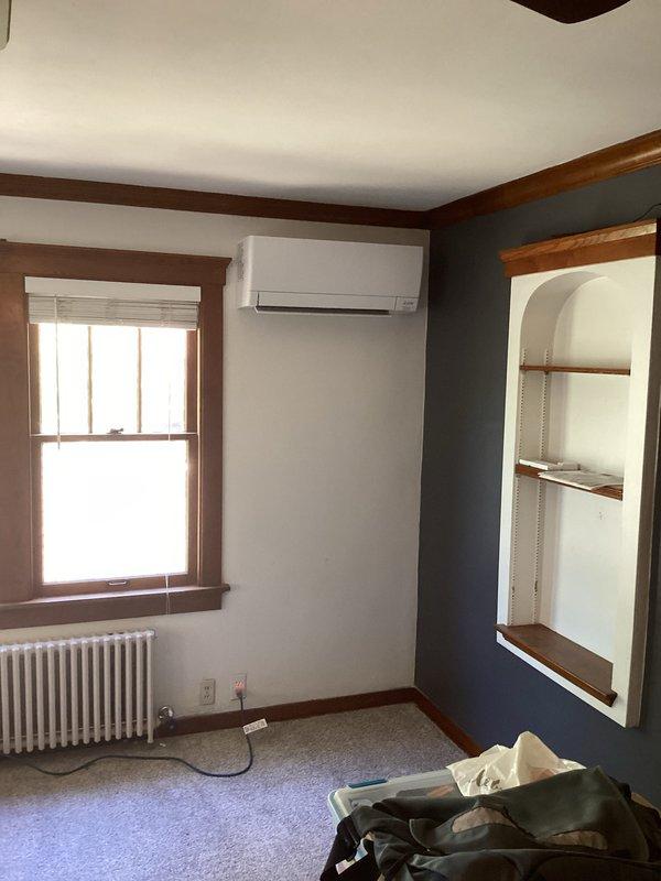 # HVAC Mini Split Installation

Completed full installation of ductless mini split air conditioning system for residential property. Mounted outdoor condenser unit on concrete pad at exterior foundation with proper clearances and secure mounting. Ran refrigerant lines, electrical connections, and condensate drain through exterior wall penetration with appropriate sealing. Installed indoor air handler unit on interior wall at optimal height for air distribution. System positioned in living space with two-tone painted walls, mounted above existing radiator heating system. Completed all electrical connections from outdoor unit to indoor evaporator, secured line set with proper insulation and protection. Tested system operation, verified refrigerant charge, confirmed proper drainage, and ensured all connections were leak-free and secure. System commissioned and left in full working order.
