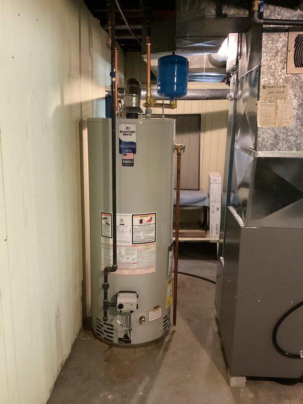 Completed residential water heater installation in basement utility room. Installed new white cylindrical water heater with proper copper piping connections, including vertical and horizontal runs with brass fittings and shut-off valves. Mounted blue thermal expansion tank to ceiling area and integrated into plumbing system per code requirements. All connections secured and tested for proper operation.
