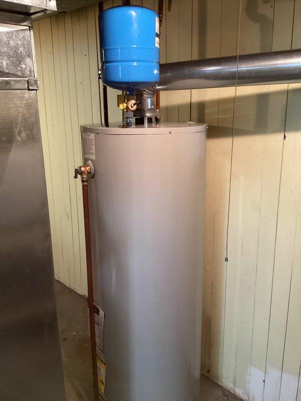 Completed residential water heater installation in basement utility room. Installed new white cylindrical water heater with proper copper piping connections, including vertical and horizontal runs with brass fittings and shut-off valves. Mounted blue thermal expansion tank to ceiling area and integrated into plumbing system per code requirements. All connections secured and tested for proper operation.
