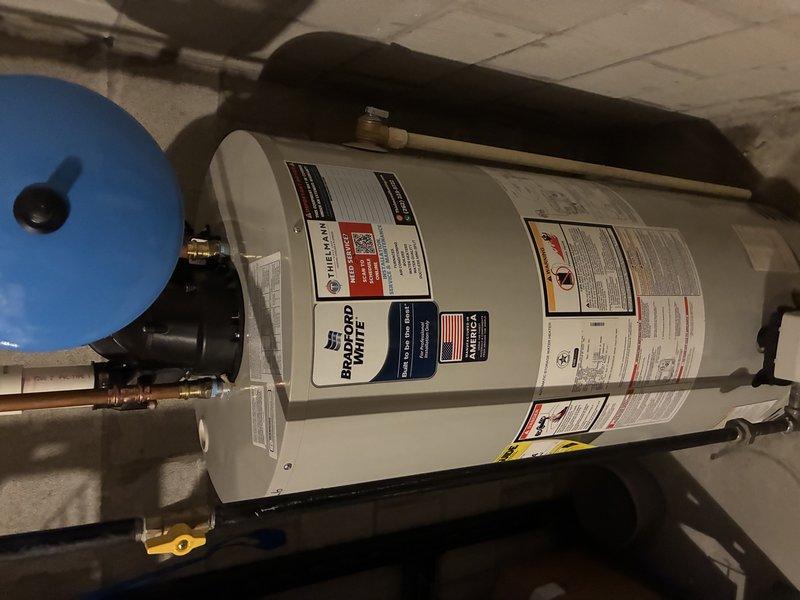 Completed installation of Bradford White tanked water heater in basement utility area. Installed new unit with proper plumbing connections including copper piping with soldered joints, brass fittings, and shut-off valve. Mounted blue expansion tank on wall and connected to system per manufacturer specifications. Verified all connections, tested system operation, and ensured compliance with plumbing codes and safety requirements.
