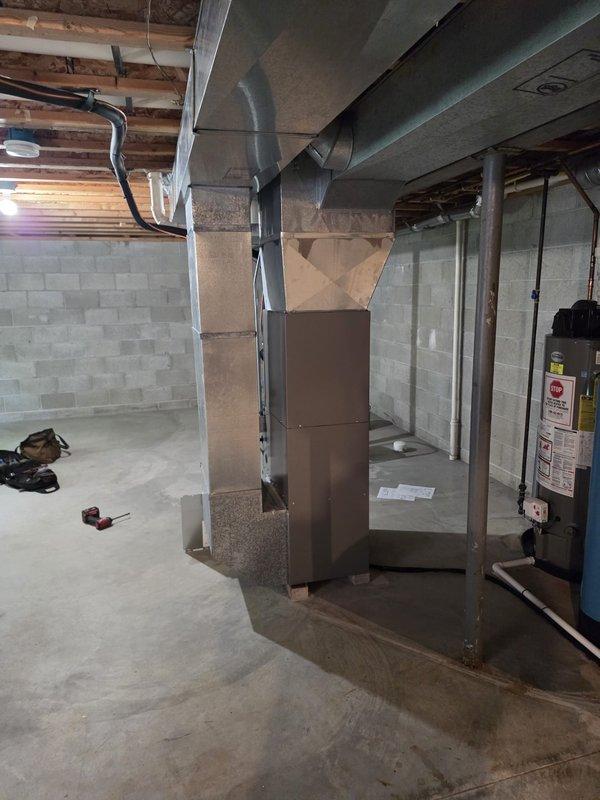 Completed full HVAC system installation including new furnace and air conditioning unit with all required permits and electrical work. Installed brown metal furnace unit in basement mechanical room with properly configured silver ductwork connecting to overhead distribution system, including both rectangular and cylindrical metal sections running along ceiling joists. Mounted new Lennox brand condenser unit on black rubber pad against exterior wall with proper refrigerant line routing and electrical disconnect, ensuring all connections meet code requirements and manufacturer specifications.