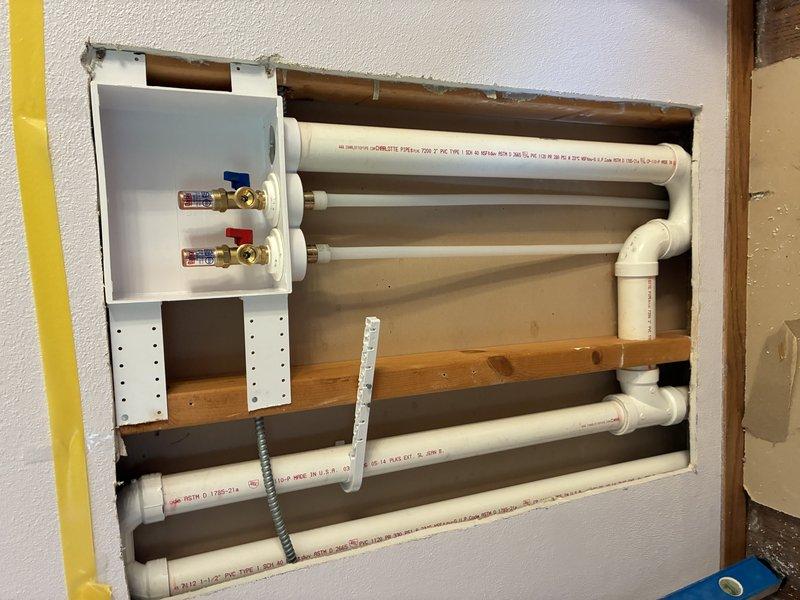 Completed laundry relocation installation with new water supply lines and drain piping. Installed hot and cold water valve assemblies with brass fittings and control handles mounted on wall bracket within framed cavity, routed PVC supply and drain lines through wall structure with proper support and securing hardware, and positioned all plumbing components to accommodate relocated laundry equipment per specifications.