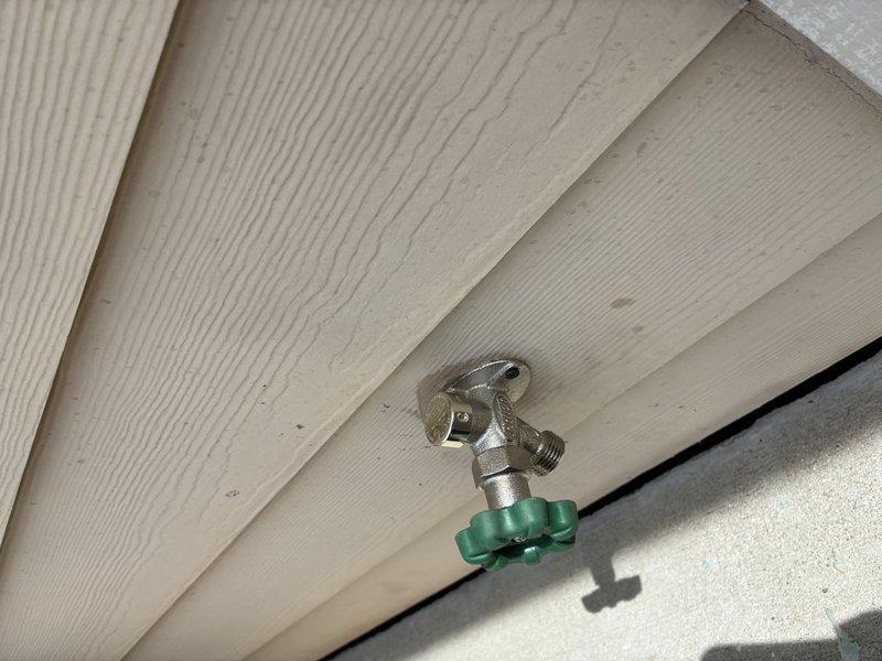 Completed installation of new exterior hose bib on residential structure, requiring access through interior wall cavity to properly connect water supply line. Mounted chrome-plated brass spigot with green handle knob on cream-colored vinyl siding, ensuring secure attachment to wall framing and proper weatherproofing of penetration point.