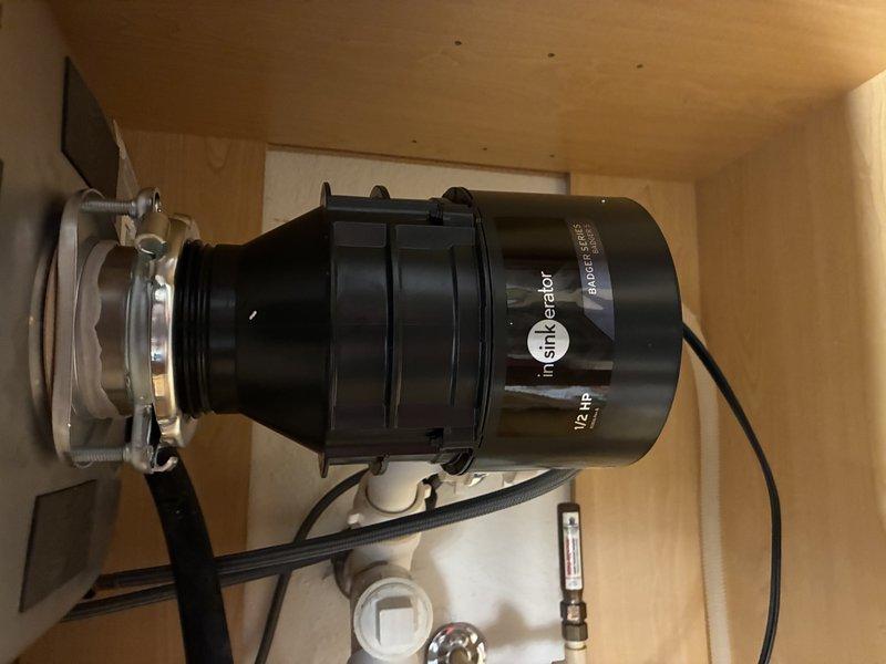 Performed diagnostic evaluation of InSinkErator garbage disposal unit. Upon inspection, determined that the existing disposal has reached end of service life and requires replacement. Identified additional concern with malfunctioning main water shut-off valve in condo unit, which will need attention to ensure proper water control for future plumbing work. Documented current disposal installation including mounting flange, electrical connections, and PVC drain configuration beneath sink. Photographed existing unit and drain assembly to provide visual record of current system setup prior to replacement work.