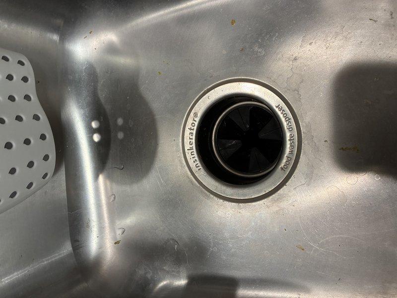 Performed diagnostic evaluation of InSinkErator garbage disposal unit. Upon inspection, determined that the existing disposal has reached end of service life and requires replacement. Identified additional concern with malfunctioning main water shut-off valve in condo unit, which will need attention to ensure proper water control for future plumbing work. Documented current disposal installation including mounting flange, electrical connections, and PVC drain configuration beneath sink. Photographed existing unit and drain assembly to provide visual record of current system setup prior to replacement work.