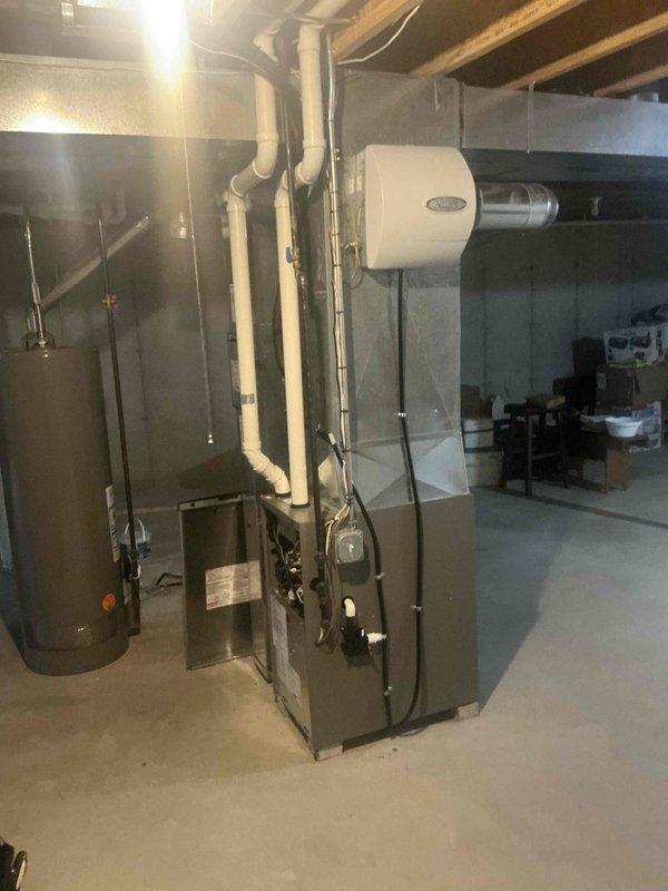 Completed HVAC equipment assessment for furnace replacement estimate in basement mechanical room. Evaluated existing dark-paneled furnace unit with blower access, associated white PVC venting system extending to ceiling joists, and existing electrical panel with dual columns of circuit breakers to determine compatibility requirements for new equipment installation. Documented current system configuration including humidifier/air treatment device mounted on ductwork, concrete floor conditions, and access parameters for replacement work.