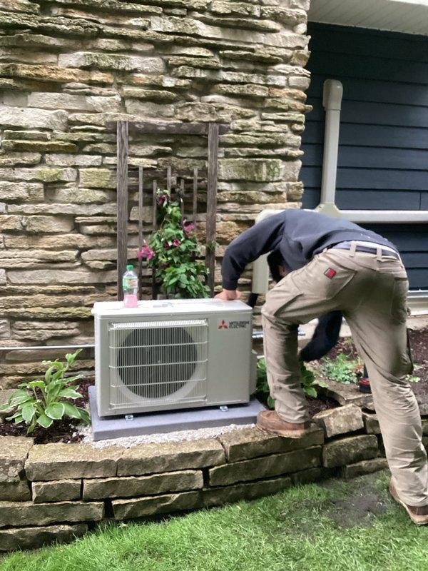 Completed installation of Mitsubishi mini-split heat pump system at residential property. Mounted outdoor condenser unit on concrete pad against stone exterior wall, installed refrigerant line set and electrical connections through exterior wall penetration, and routed condensate drainage via white PVC piping along building exterior. Indoor air handler installation included proper refrigerant charging, system commissioning, and operational testing to verify heating and cooling performance. All work performed in accordance with manufacturer specifications and local permit requirements, with licensed electrician completing final electrical connections.