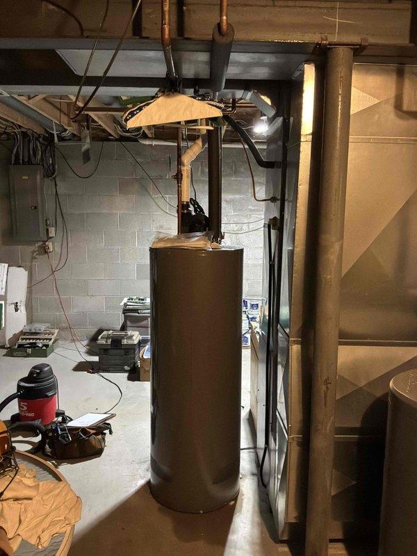 Responded to emergency water heater replacement request due to active water leak beneath existing unit. Assessed brown cylindrical water heater in basement utility area with visible water accumulation at base, confirmed immediate replacement necessary to prevent further water damage and restore hot water service. Documented existing installation including copper supply lines, black rubber connections, proximity to water softener system, and current venting configuration through exposed ceiling joists for replacement estimate and planning purposes.
