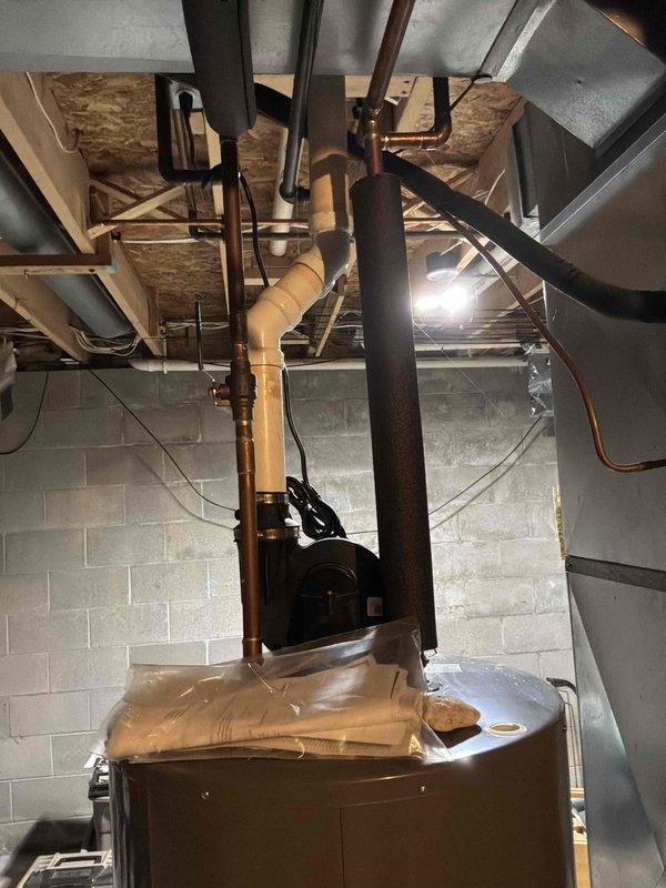 Responded to emergency water heater replacement request due to active water leak beneath existing unit. Assessed brown cylindrical water heater in basement utility area with visible water accumulation at base, confirmed immediate replacement necessary to prevent further water damage and restore hot water service. Documented existing installation including copper supply lines, black rubber connections, proximity to water softener system, and current venting configuration through exposed ceiling joists for replacement estimate and planning purposes.