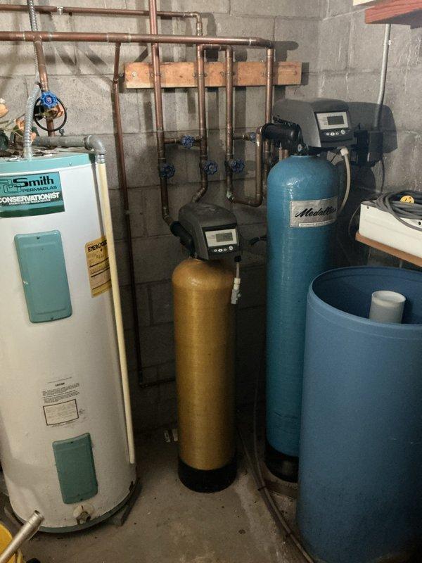 Responded to service call for non-operational residential water heater at small congregation facility with minimal hot water usage. Inspected basement mechanical room containing Smith tanked water heater and associated water treatment system including iron filtration equipment (iron curtain). Performed visual assessment of water heater operation, verified proper function of heating elements and thermostat controls. Evaluated water treatment system consisting of multiple cylindrical treatment tanks with digital control heads and blue ball valve shutoff system on copper supply lines. Confirmed pressure gauge readings within normal parameters. Assessed iron filtration system operation to ensure proper water conditioning for water heater longevity. System found operational with no immediate repairs required at time of inspection.