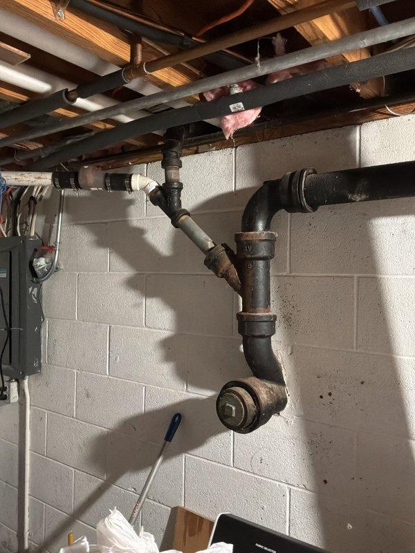 Conducted comprehensive assessment of existing sewer stack system in basement area. Inspected severely corroded cast iron drain pipes showing extensive rust deterioration throughout multiple joints, elbows, and connection points. The existing piping configuration exhibited significant age-related degradation with reddish-brown corrosion compromising structural integrity of the drainage system. 

Documented current plumbing infrastructure including horizontal and vertical pipe runs mounted between floor joists, noting multiple right-angle connections and elbow fittings in deteriorated condition. Evaluated replacement scope requiring removal of compromised cast iron stack and installation of new PVC drainage piping system to restore proper waste water flow and prevent potential leaks or failures.