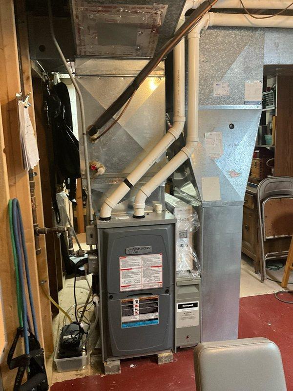 Completed HVAC estimate for furnace replacement in basement location. Inspected existing dark gray residential furnace unit with associated ductwork, white PVC venting system, and integrated humidifier mounted to supply duct. Documented current system configuration including furnace positioning on wooden platform, metal plenum connections, condensate drain lines, and beige humidifier unit with electrical connections. Photographed equipment specifications, manufacturer labels, and overall mechanical room layout to provide accurate replacement proposal for aging heating system.