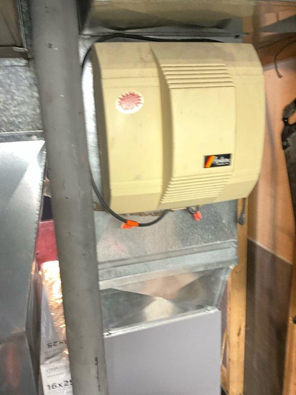 Completed HVAC estimate for furnace replacement in basement location. Inspected existing dark gray residential furnace unit with associated ductwork, white PVC venting system, and integrated humidifier mounted to supply duct. Documented current system configuration including furnace positioning on wooden platform, metal plenum connections, condensate drain lines, and beige humidifier unit with electrical connections. Photographed equipment specifications, manufacturer labels, and overall mechanical room layout to provide accurate replacement proposal for aging heating system.
