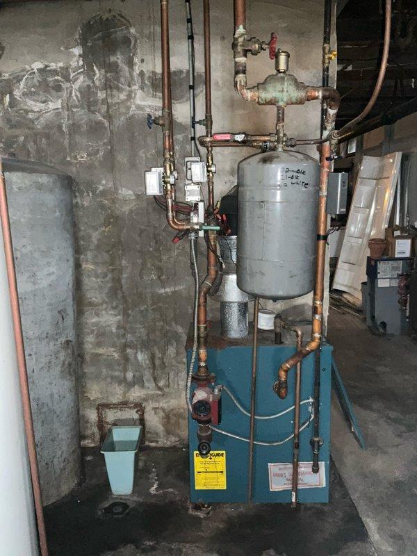 Conducted on-site evaluation for natural gas boiler replacement in basement mechanical room. Current boiler system, mounted on blue metal base with gray expansion tank and extensive copper piping network, requires significant repairs that make replacement more cost-effective than restoration. Documented existing equipment configuration including expansion tank marked "2-4½", multiple control valves with red handles, electrical junction boxes, and interconnected plumbing assembly. Assessed deteriorated concrete block walls showing water staining and age-related damage. Photographed complete system layout including water heater, overhead joists, piping runs, brass fittings, and floor-mounted components to provide accurate replacement specifications and installation planning.