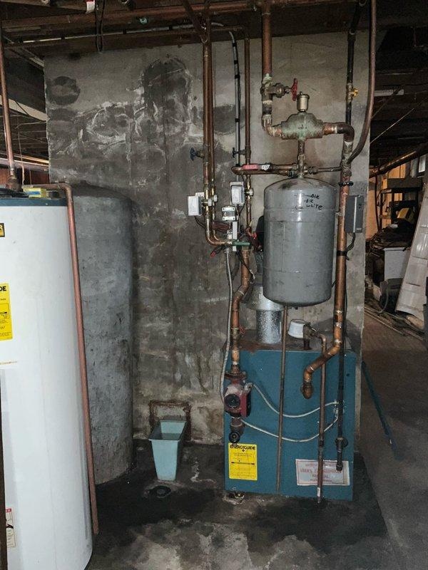 Conducted on-site evaluation for natural gas boiler replacement in basement mechanical room. Current boiler system, mounted on blue metal base with gray expansion tank and extensive copper piping network, requires significant repairs that make replacement more cost-effective than restoration. Documented existing equipment configuration including expansion tank marked "2-4½", multiple control valves with red handles, electrical junction boxes, and interconnected plumbing assembly. Assessed deteriorated concrete block walls showing water staining and age-related damage. Photographed complete system layout including water heater, overhead joists, piping runs, brass fittings, and floor-mounted components to provide accurate replacement specifications and installation planning.
