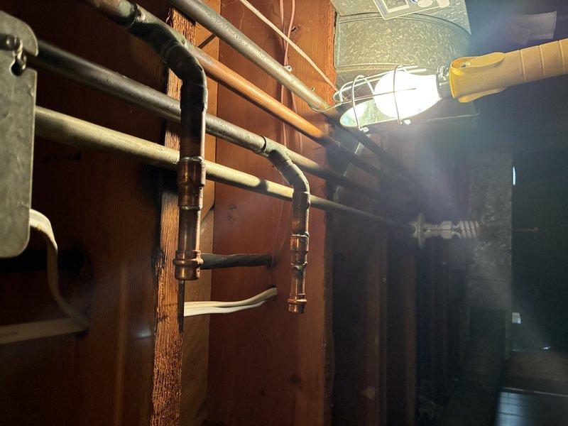 Responded to emergency call for damaged copper L-pipe resulting from failed homeowner repair attempt. Upon arrival, assessed the compromised piping system in the attic/ceiling space where copper refrigerant or plumbing lines run parallel between exposed wooden rafters. Documented existing conditions showing multiple copper pipes with tan insulation tape sections and mounting brackets securing lines to structural beams. 

Evaluated secondary piping infrastructure in basement utility area where dark insulated pipes run horizontally along exposed brick walls, secured with metal mounting brackets at joints and directional changes. Completed diagnostic inspection of both affected areas to determine extent of damage and necessary repairs to restore proper function and prevent further water damage or system failure.