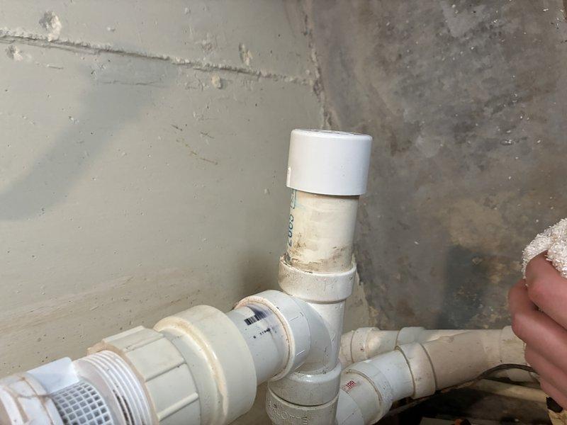 Responded to service call for reported water leak in basement utility area. Upon arrival, performed diagnostic inspection of plumbing assembly consisting of white PVC piping with multiple fittings and connections arranged vertically along corner wall. Identified older-style valve component at base of assembly showing significant age-related deterioration, including visible corrosion, rust-colored staining, and discoloration around circular face and body. Examined PVC pipe connections, threaded fittings, and flow meter device with monitoring equipment. Inspected junction points where vertical pipe connects to elbow joint and horizontal runs, noting wear patterns and residue accumulation around connection points. Documented condition of plumbing system through photographic evidence for estimate preparation. Leak source assessment completed, findings indicate compromised valve assembly requiring replacement to resolve water intrusion issue.