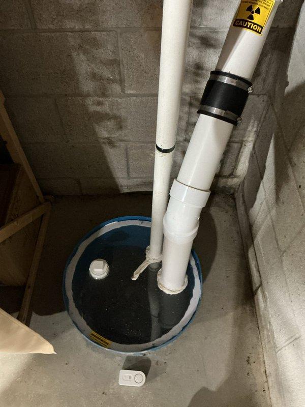 Conducted on-site evaluation of existing sump pump system to provide estimate for backup pump installation. Assessed current setup consisting of primary sump pump with vertical PVC discharge piping secured to concrete block wall with metal clamps, discharging into circular sump pit with blue rim. Evaluated exterior drainage system including white PVC and black corrugated flexible piping extending from foundation to direct water away from structure. Discussed options for secondary backup system including battery-powered backup pump and water-powered backup pump alternatives to ensure continuous operation during power outages or primary pump failure. Documented existing conditions and drainage infrastructure for estimate preparation.