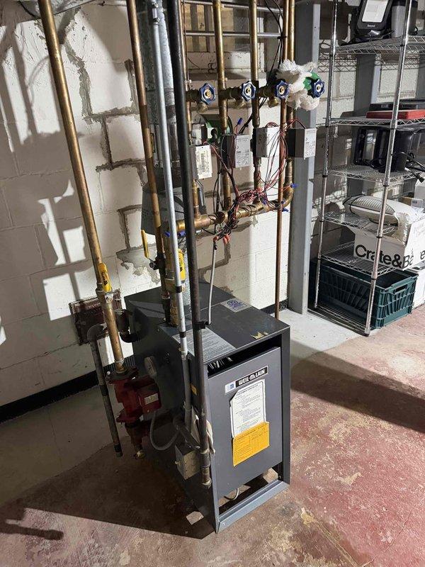 Conducted comprehensive assessment of existing residential boiler system for replacement estimate. Evaluated current gray cabinet-style boiler unit with manifold distribution system featuring multiple zone valves and copper supply lines. Documented existing installation including piping configuration, manifold assembly with blue and green-handled zone valves, and associated circulation pump. Assessed condition of copper piping network, insulated supply lines, and connection points throughout the heating distribution system. Photographed current equipment specifications, manufacturer labels, and existing plumbing arrangement to provide accurate replacement recommendations and ensure proper system sizing for the application.