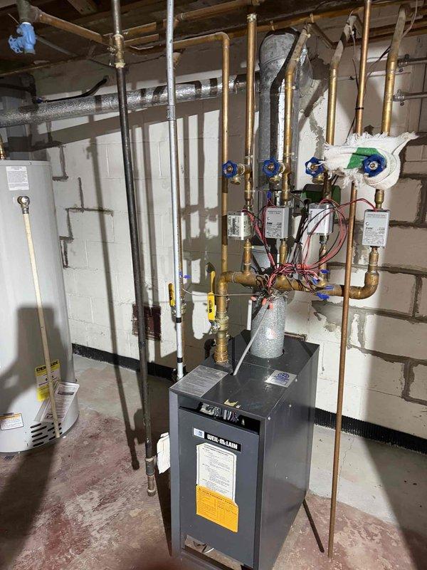 Conducted comprehensive assessment of existing residential boiler system for replacement estimate. Evaluated current gray cabinet-style boiler unit with manifold distribution system featuring multiple zone valves and copper supply lines. Documented existing installation including piping configuration, manifold assembly with blue and green-handled zone valves, and associated circulation pump. Assessed condition of copper piping network, insulated supply lines, and connection points throughout the heating distribution system. Photographed current equipment specifications, manufacturer labels, and existing plumbing arrangement to provide accurate replacement recommendations and ensure proper system sizing for the application.