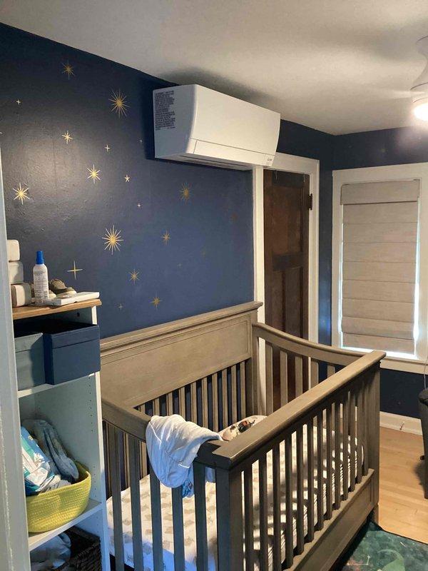 Completed full installation of Mitsubishi ductless mini-split system in nursery room with outdoor condenser unit mounted on exterior brick wall. Installation included proper permitting, electrical work by licensed electrician, refrigerant line set installation with exterior conduit protection, indoor wall-mounted air handler placement above doorway, and decorative stone gravel base around outdoor unit for drainage and aesthetic appeal.