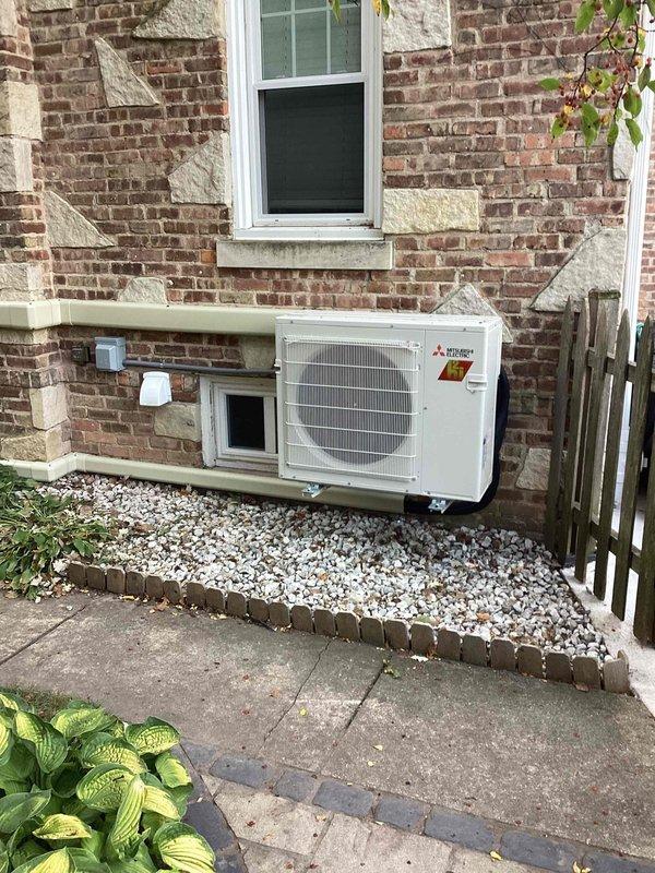 Completed full installation of Mitsubishi ductless mini-split system in nursery room with outdoor condenser unit mounted on exterior brick wall. Installation included proper permitting, electrical work by licensed electrician, refrigerant line set installation with exterior conduit protection, indoor wall-mounted air handler placement above doorway, and decorative stone gravel base around outdoor unit for drainage and aesthetic appeal.