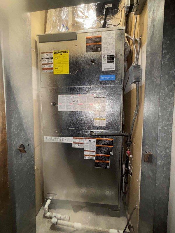 Completed full Magicpak HVAC system installation in basement utility area. Unit was transported via stairs using dolly due to non-operational elevator, requiring additional crew member for safe handling. Installed vertical furnace unit with connecting galvanized metal ductwork on both sides, routed white PVC condensate drainage lines along floor, and connected flexible gas line with proper wall mounting. Mounted thermostat control with cooling, heating, and fan operation settings, and left manufacturer documentation and components with installation. All equipment tested and operational upon completion.