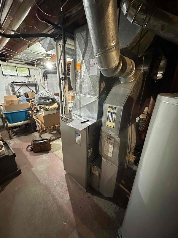 Conducted comprehensive HVAC system evaluation for furnace and air conditioning replacement estimate. Assessed existing gray furnace unit in basement utility area, including ductwork connections and overall system condition, and inspected outdoor Carrier brand AC condenser unit positioned on concrete pad with electrical connections to determine replacement specifications and requirements.