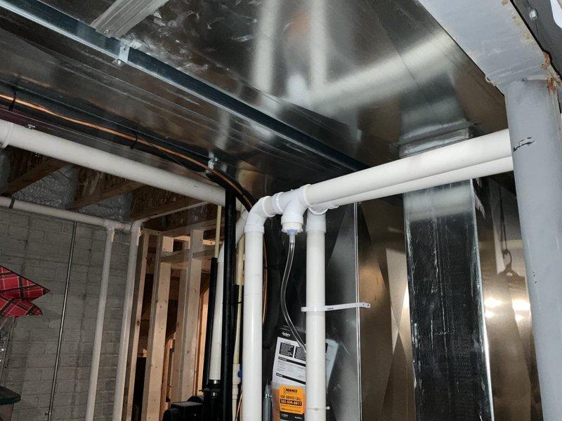 Conducted site assessment for basement buildout HVAC ductwork installation. Documented existing mechanical infrastructure including large rectangular sheet metal ductwork with visible seams and joints, white PVC drain lines with elbow joints, and various copper and black piping systems secured with mounting straps. Photographed exposed wooden framing, concrete block walls, and ceiling joists to evaluate routing options for new duct runs. Assessed clearances around existing galvanized ductwork with angular V-shaped configuration and verified compatibility with planned basement finishing work.