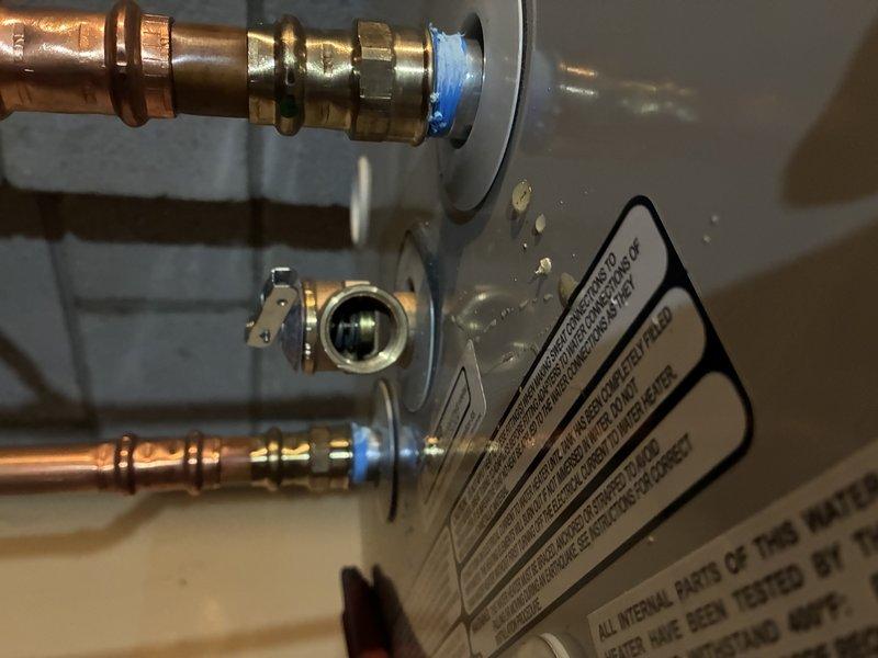 Responded to service call for water heater leaking from pipe connections. Upon arrival, inspected tanked water heater and identified active leak at copper pipe compression fittings with blue plastic ferrules. Evidence of significant water damage was visible on floor surrounding unit, with extensive staining and discoloration indicating ongoing moisture issues. Assessed plumbing connections at top of water heater, noting corrosion on copper pipes and mineral deposits around connection points. Tightened compression fittings and verified proper seal at both inlet and outlet connections. Cleaned area and monitored for additional leakage to confirm repair effectiveness.