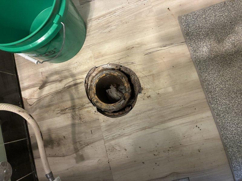 Assessed a failed cast iron toilet flange showing heavy corrosion, rust scaling, and broken flange ring sections, with moisture-related deterioration of the surrounding subfloor and corroded cast iron drain piping visible in the underfloor area. Prepared an estimate to remove approximately 7–8 feet of deteriorated cast iron drain piping and replace it with PVC, including a new toilet flange, and to include the bathtub drain in the same scope of work.