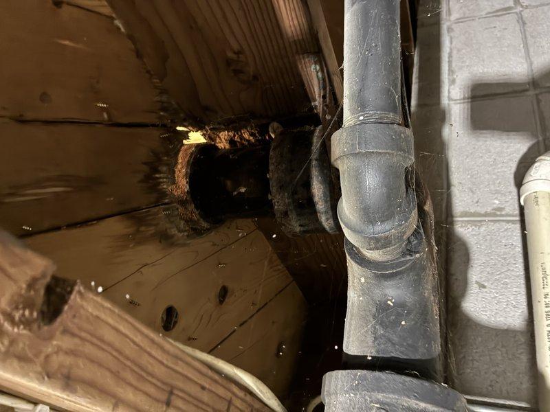 Assessed a failed cast iron toilet flange showing heavy corrosion, rust scaling, and broken flange ring sections, with moisture-related deterioration of the surrounding subfloor and corroded cast iron drain piping visible in the underfloor area. Prepared an estimate to remove approximately 7–8 feet of deteriorated cast iron drain piping and replace it with PVC, including a new toilet flange, and to include the bathtub drain in the same scope of work.