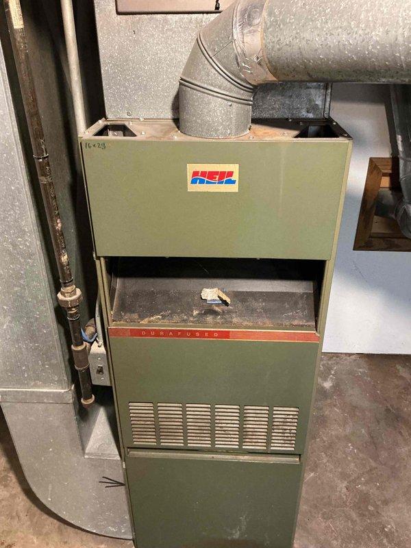Performed a visual inspection of an aged Heil upright gas furnace to evaluate its serviceability, noting surface rust and heavy dust accumulation at the flue/duct collar and burner compartment, along with debris on the interior ledge. Gas piping, shutoff valve, and flue connections were observed and documented, and the unit's age and condition were assessed for continued use.
