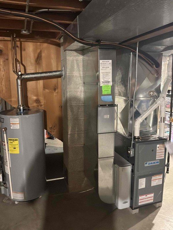 Performed an indoor air quality (IAQ) estimate, inspecting the Daikin gas furnace, galvanized supply plenum and ductwork, PVC venting, and the atmospherically vented water heater in the basement mechanical area. Equipment and ductwork appeared clean and in good condition with no visible rust, frost, or damage, and an existing cabinet-style device (air cleaner/humidifier) was noted on the supply duct. Documented the system with photos to evaluate IAQ improvement options.