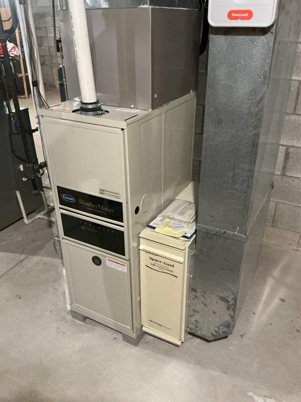 Conducted an on-site evaluation for replacement of an existing Carrier WeatherMaker Infinity high-efficiency natural gas furnace located in the basement mechanical area, with an associated Space-Gard air cleaner and galvanized supply trunk. Inspected the PVC vent and condensate piping, noting damp concrete flooring and staining near the condensate drain run and adjacent water treatment tank, and documented existing equipment condition with photos to support the replacement proposal.