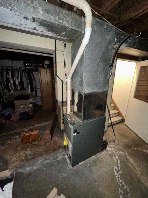 Responded to a non-operational furnace and performed an on-site evaluation of the existing upright gas furnace, plenum, and ductwork in the basement utility area to prepare a replacement estimate. Documented the unit's condition, including the dusty, aged supply plenum and ductwork, the condensate/PVC piping, and the stubbed PVC and corroded metal piping near the base, noting the stained concrete floor and clearance around the equipment for installation planning. Replacement recommendations were prepared based on the findings.