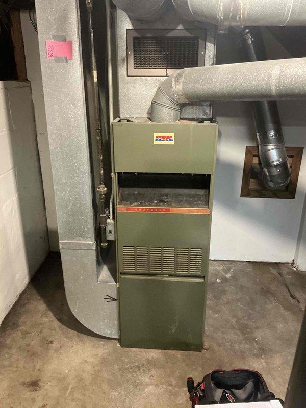 Performed an on-site replacement estimate for a 62-year-old Heil 80% AFUE natural gas upflow furnace, documenting the unit's condition, nameplate data, and surrounding galvanized supply/return ductwork and flue venting through the wall. Noted significant dust and debris accumulation inside the cabinet, aged and soiled equipment, and a stained, uneven concrete floor at the base of the unit. Documented the existing configuration to support sizing and replacement recommendations for a new high-efficiency furnace.