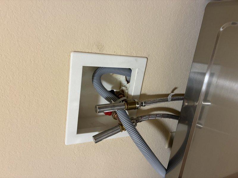 Inspected the laundry wall outlet box and diagnosed a leak at the cold water supply valve and hose connection, which prevented installation of the new washer and dryer. Evaluated the valve assembly, braided stainless supply hoses, and drain hose, noting staining and corrosion on the box interior and surrounding wall consistent with prolonged moisture exposure. Documented the condition with photographs for repair planning.