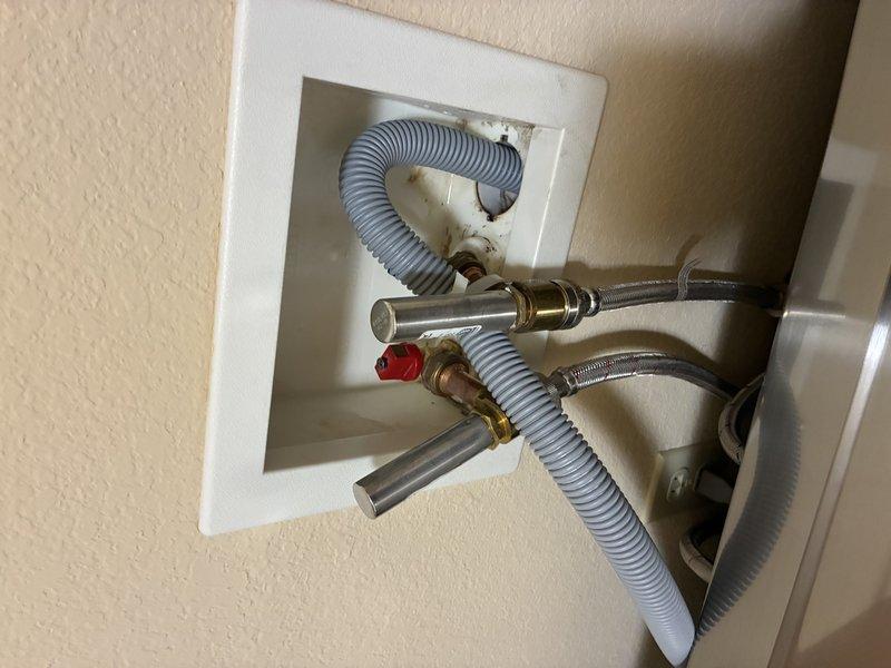 Inspected the laundry wall outlet box and diagnosed a leak at the cold water supply valve and hose connection, which prevented installation of the new washer and dryer. Evaluated the valve assembly, braided stainless supply hoses, and drain hose, noting staining and corrosion on the box interior and surrounding wall consistent with prolonged moisture exposure. Documented the condition with photographs for repair planning.