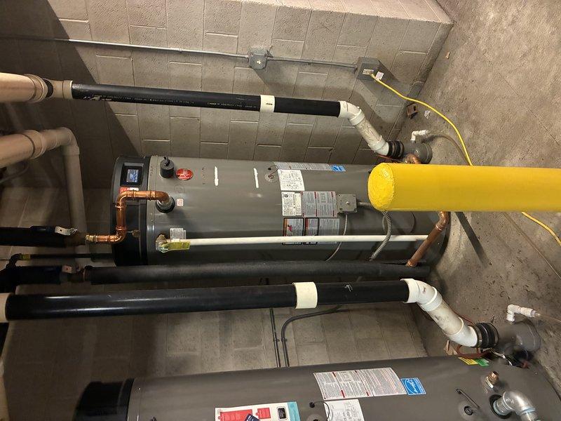 Inspected two Rheem commercial tank-type water heaters reporting fault codes, verifying the digital control displays, gas control valves, electrical connections, and associated copper and PVC piping. Equipment appeared intact with no frost or rust on the units, though a brown/orange mineral stain was noted on the concrete beneath the pipe fittings, indicating possible minor leakage or corrosion at the connections that should be monitored.