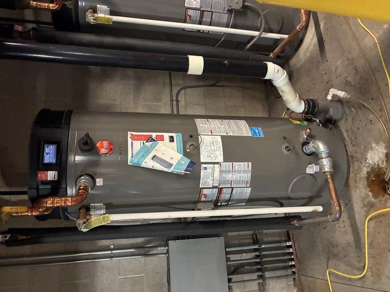 Inspected two Rheem commercial tank-type water heaters reporting fault codes, verifying the digital control displays, gas control valves, electrical connections, and associated copper and PVC piping. Equipment appeared intact with no frost or rust on the units, though a brown/orange mineral stain was noted on the concrete beneath the pipe fittings, indicating possible minor leakage or corrosion at the connections that should be monitored.