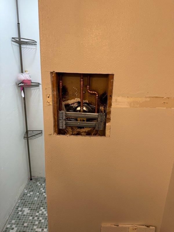 Performed diagnostic evaluation of bathroom shower system and accessed plumbing infrastructure through wall cavity. Inspected thermostatic shower valve assembly with chrome finish, verified proper installation of dual flexible metal supply hoses, and documented existing fixture configuration. Opened wall access point to examine copper supply lines, associated mechanical systems, and structural components to assess scope of work outlined in estimate.