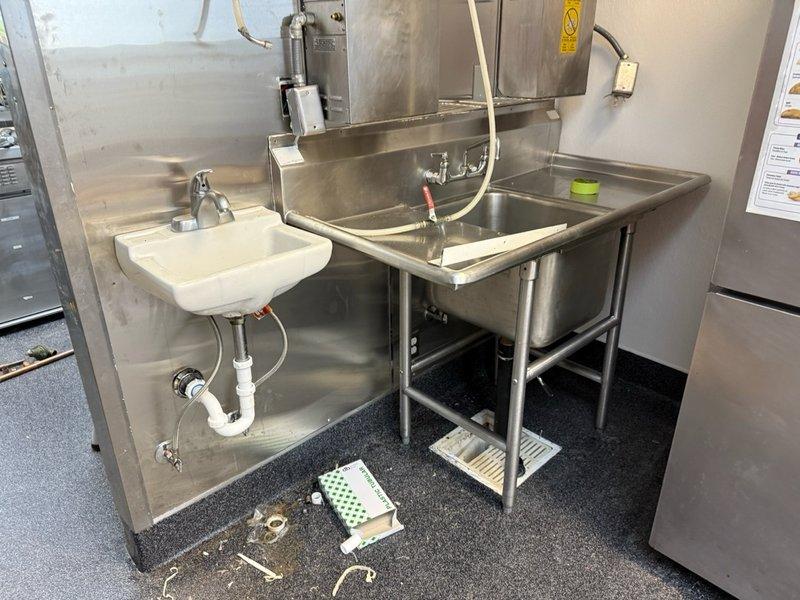 Responded to diagnostic service call for commercial kitchen plumbing evaluation. Upon arrival, inspected two-compartment stainless steel utility sink with wall-mounted hand washing station, noting visible PVC drain connections and wall-mounted spray hose assembly. Examined floor-level plumbing components including scattered brass fittings and white perforated floor drain cover positioned near sink area. Assessed secondary service area where blue rubber supply line connects to wall-mounted fitting with metallic elbow connector approximately twelve inches above floor level. Documented existing plumbing configuration and equipment conditions through photographic records for future reference and scope determination. All observations recorded for further evaluation and repair recommendations.