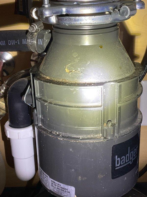 Responded to report of slow leak beneath kitchen sink. Upon inspection, identified source as InSinkErator Badger garbage disposal unit, approximately 7 years old. Leak detected at upper mounting flange assembly where disposal connects to sink drain. Observed moisture accumulation and corrosion around metal mounting ring and rubber gasket seal. Dishwasher drain connection and PVC drain piping inspected and found to be secure with no signs of leakage. Documented conditions with photographs showing wear patterns on mounting hardware and deterioration of sealing components. Provided homeowner with diagnostic findings and repair options for addressing the compromised flange seal.