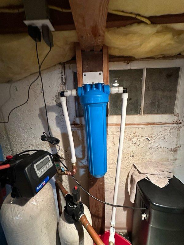 Completed diagnostic assessment of residential water filtration system. Inspected blue cylindrical water filter housing mounted to structural support, evaluated condition of copper and PVC plumbing connections, brass fittings, and associated pressure tank. Documented existing equipment configuration including water treatment control unit, valve assemblies, and piping layout for filter replacement planning.