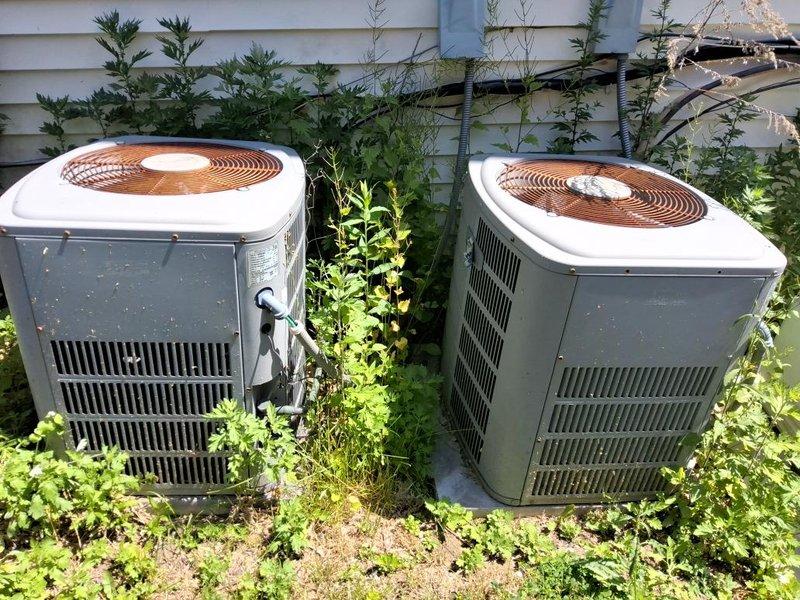 Completed estimate for residential air conditioning system. Inspected attic-mounted air handler with associated ductwork and refrigerant lines, confirming standard installation with fiberglass insulation present. Evaluated two outdoor condenser units positioned side by side with vegetation growth noted around equipment. Documented system configuration and current condition through photographic records for estimate preparation.
